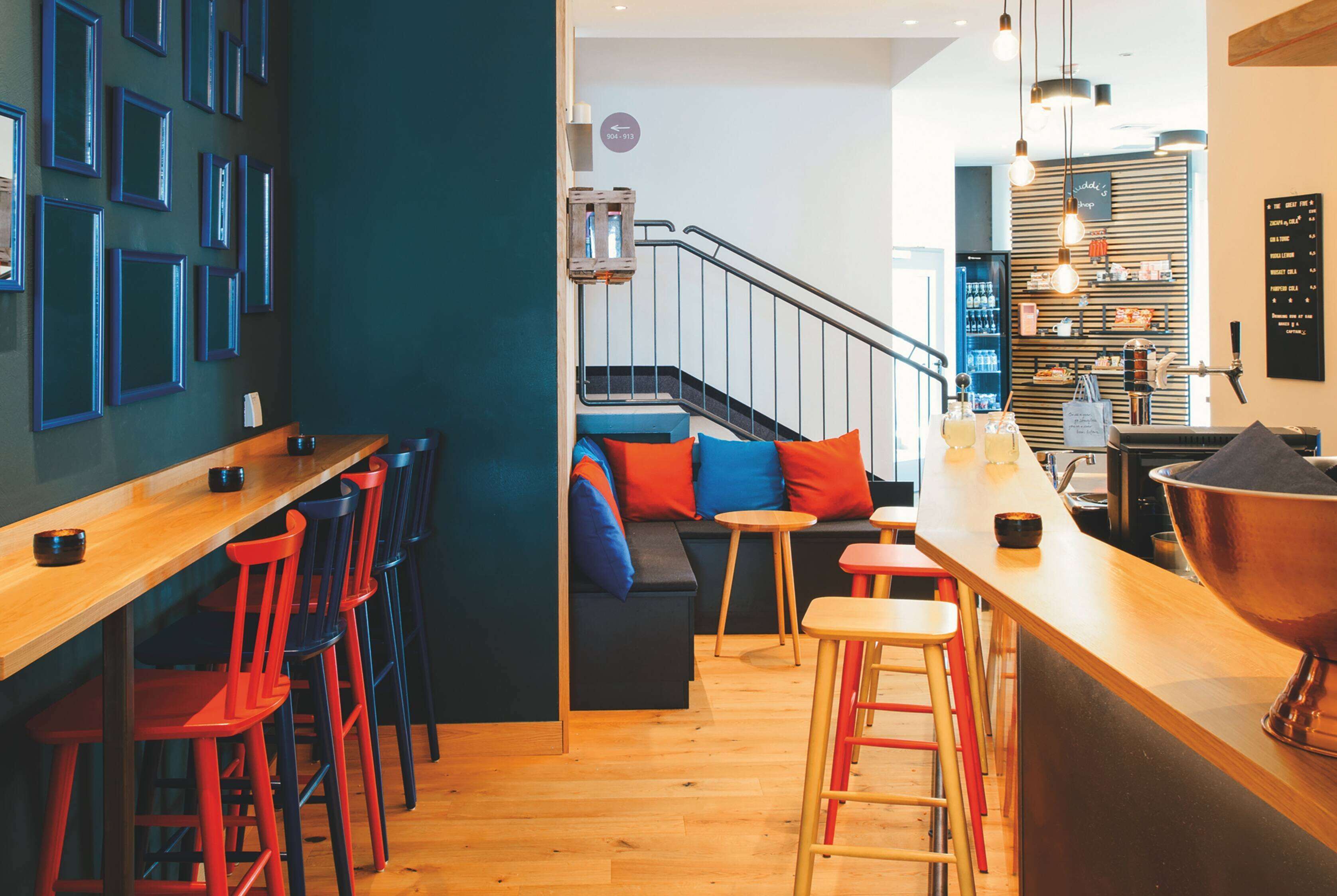 Zona de bar moderna con mostradores de madera, taburetes de bar rojos y azules, y un área de asientos con cojines de colores.