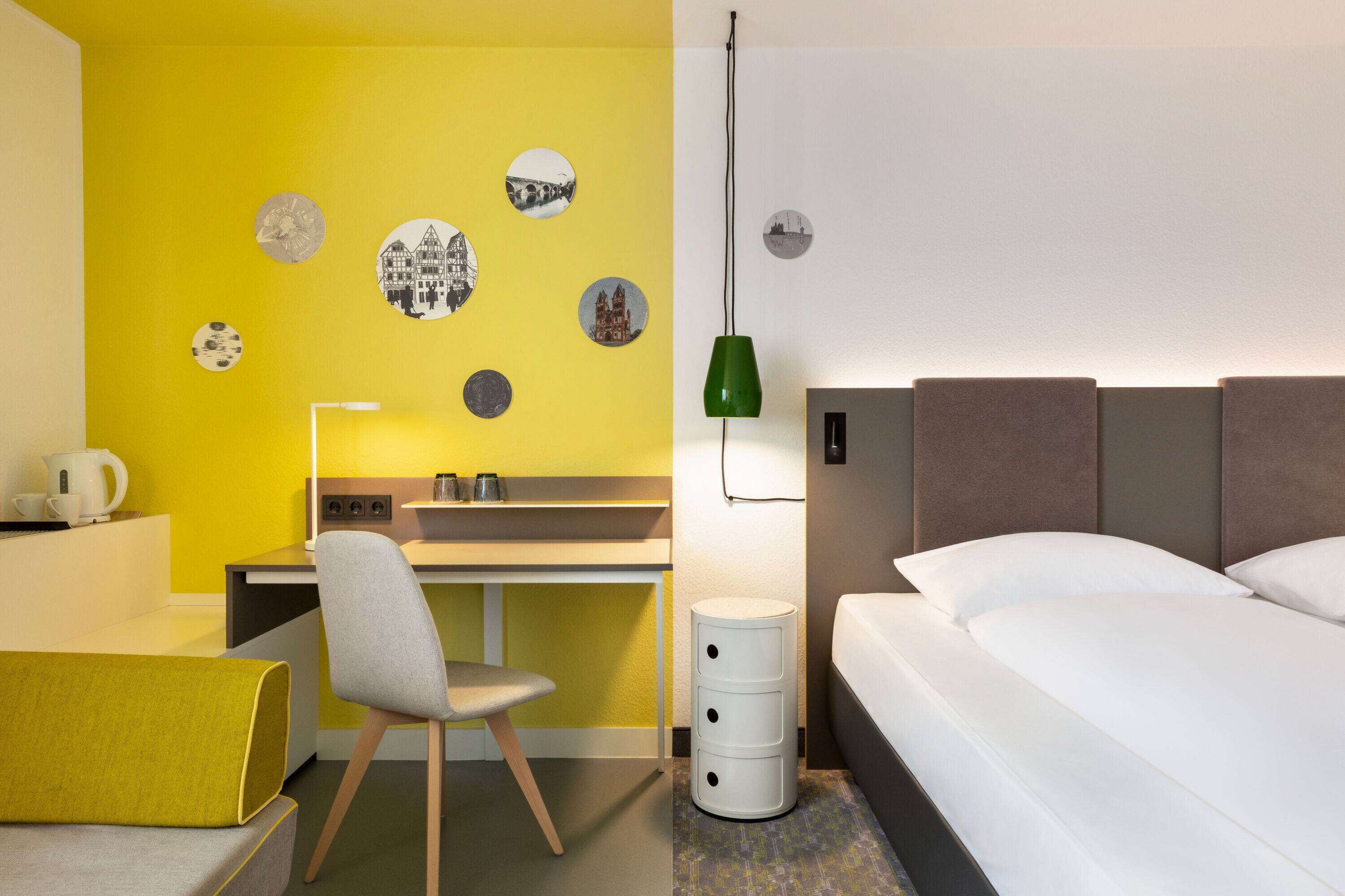 Quarto de hotel com uma parede amarela de destaque, uma cama, uma escrivaninha com cadeira, uma luminária e uma lixeira.