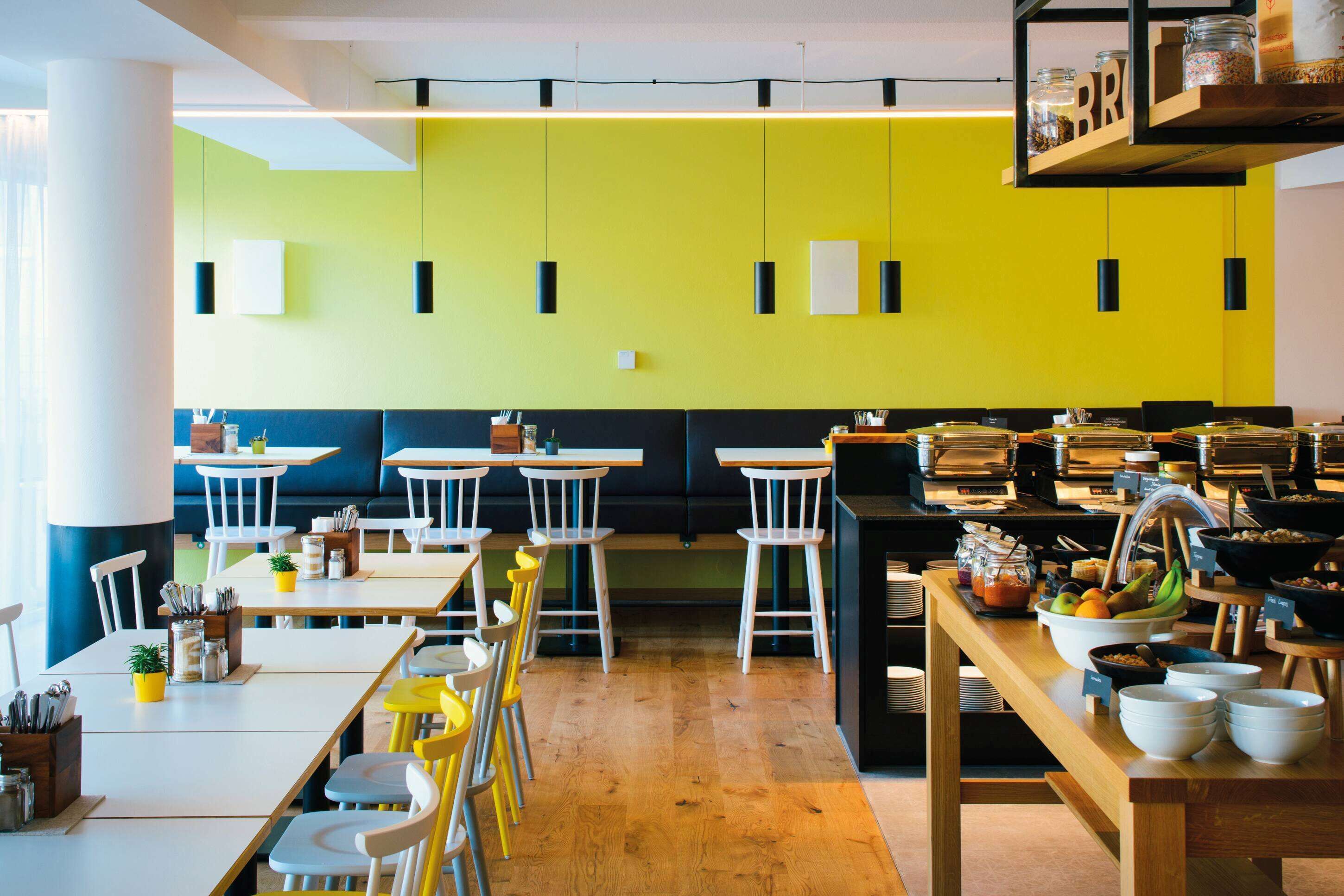 A bright dining area with yellow walls, white tables, and yellow chairs. There are shelves with jars and a buffet table with bowls and fruits.