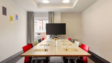 Un salón de conferencias con una mesa grande, sillas rojas, un televisor de pantalla plana y una ventana con cortinas.