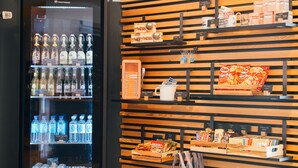 Een automaat gevuld met drankjes en snacks, een houten wand met planken waarop verschillende producten staan, en een bordje met de tekst "Muddi's Shop".