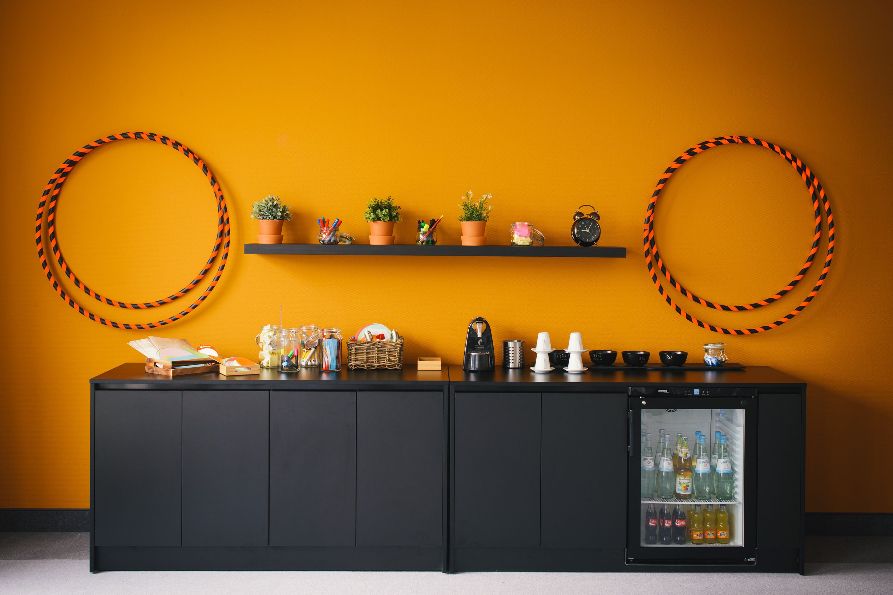 A bar area with a black cabinet, a mini-fridge stocked with beverages, and shelves with potted plants and kitchenware.