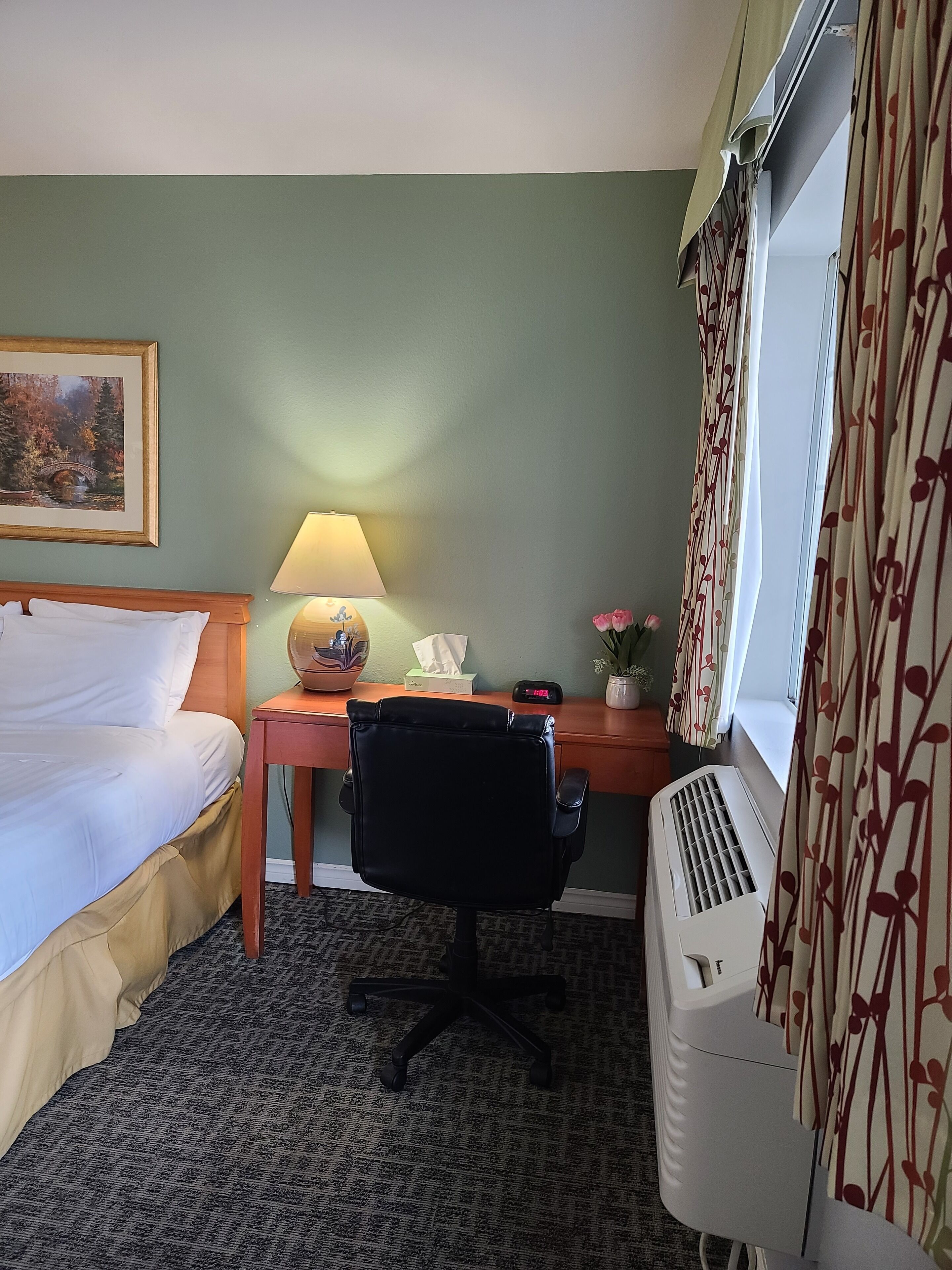 Habitación de hotel con cama, escritorio, silla, lámpara, aire acondicionado y ventana con cortinas.