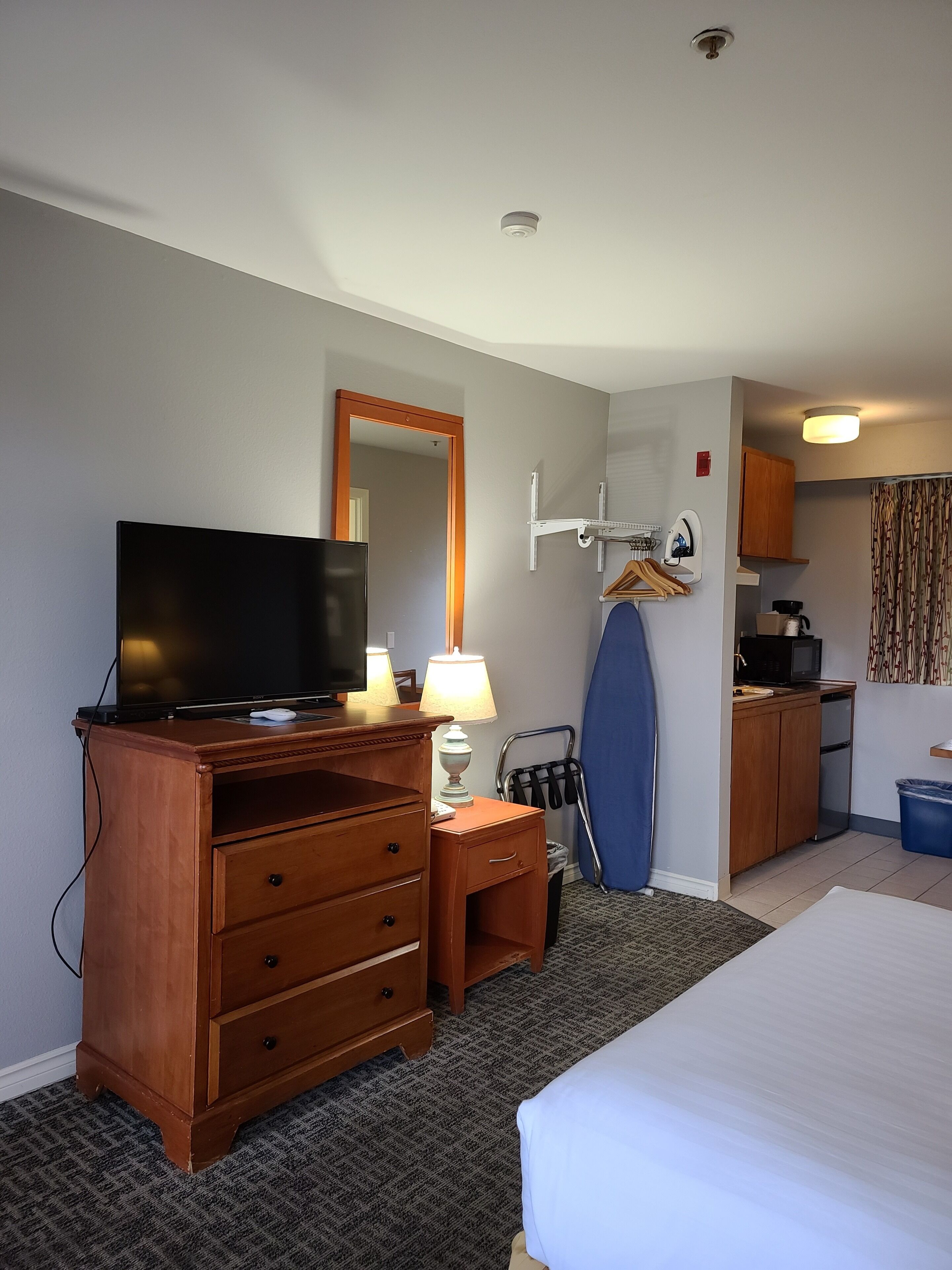 A hotel room with a bed, a wooden dresser with a TV, a desk with a lamp, a mirror, a coat rack, a kitchenette, and a shower area.