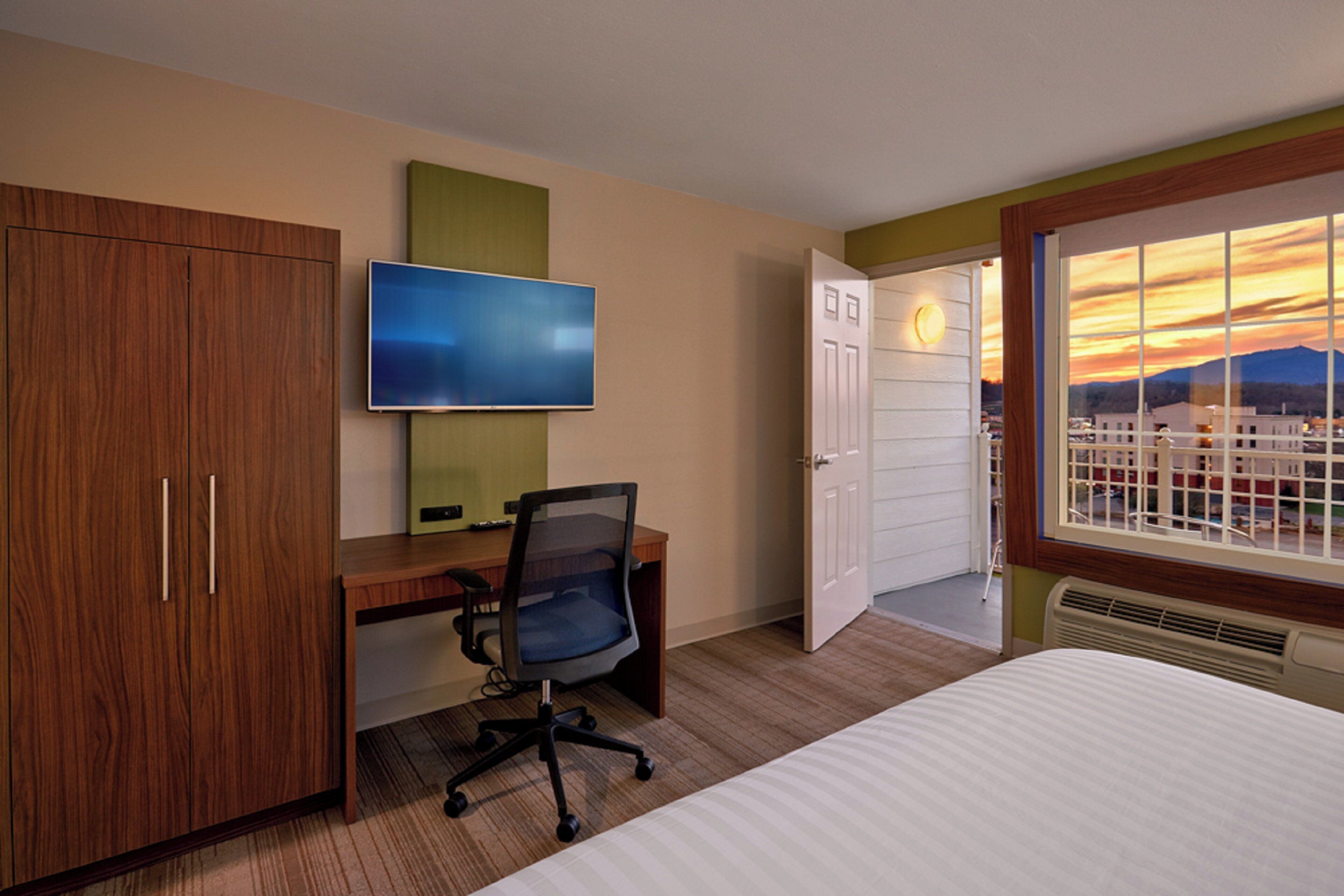 Une chambre d’hôtel avec un lit, un bureau équipé d’un ordinateur, une télévision et une fenêtre donnant sur un paysage au coucher du soleil.