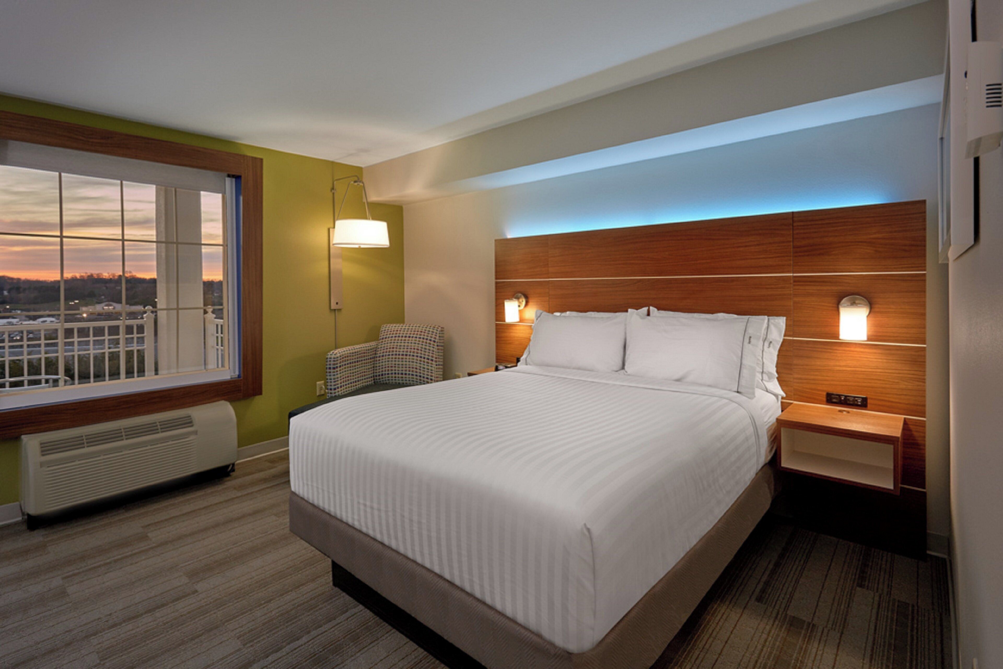 A hotel room with a large bed, a nightstand, a lamp, and a view of the city through a window.