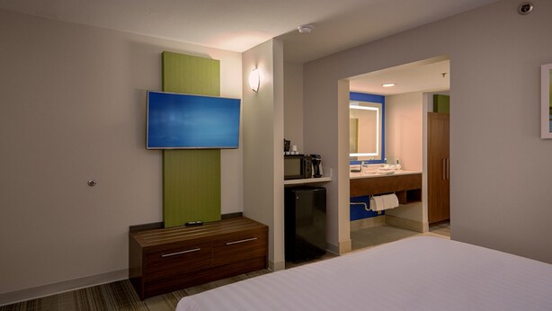 Une chambre d’hôtel avec un lit, un téléviseur fixé au mur, un petit bureau avec des tiroirs et une salle de bain visible à travers une porte ouverte.