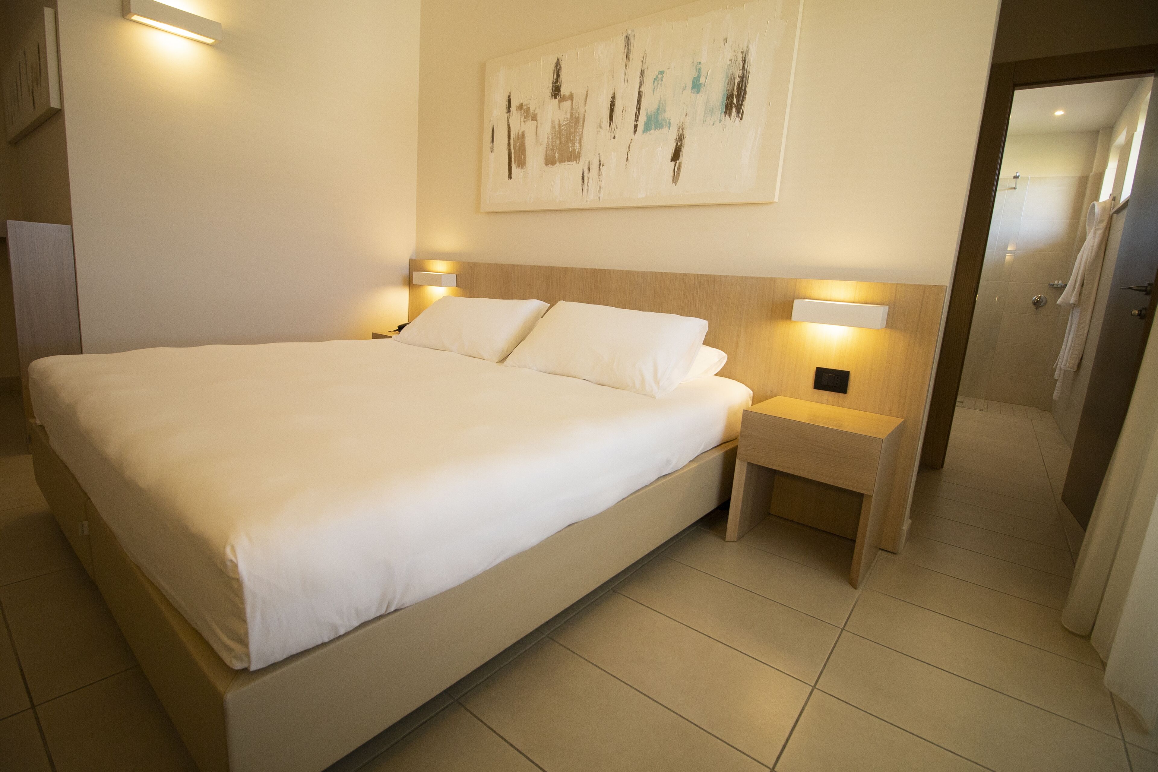 Una cama grande con sábanas blancas, dos mesitas de noche con lámparas, un cabecero de madera y un cuadro en la pared.