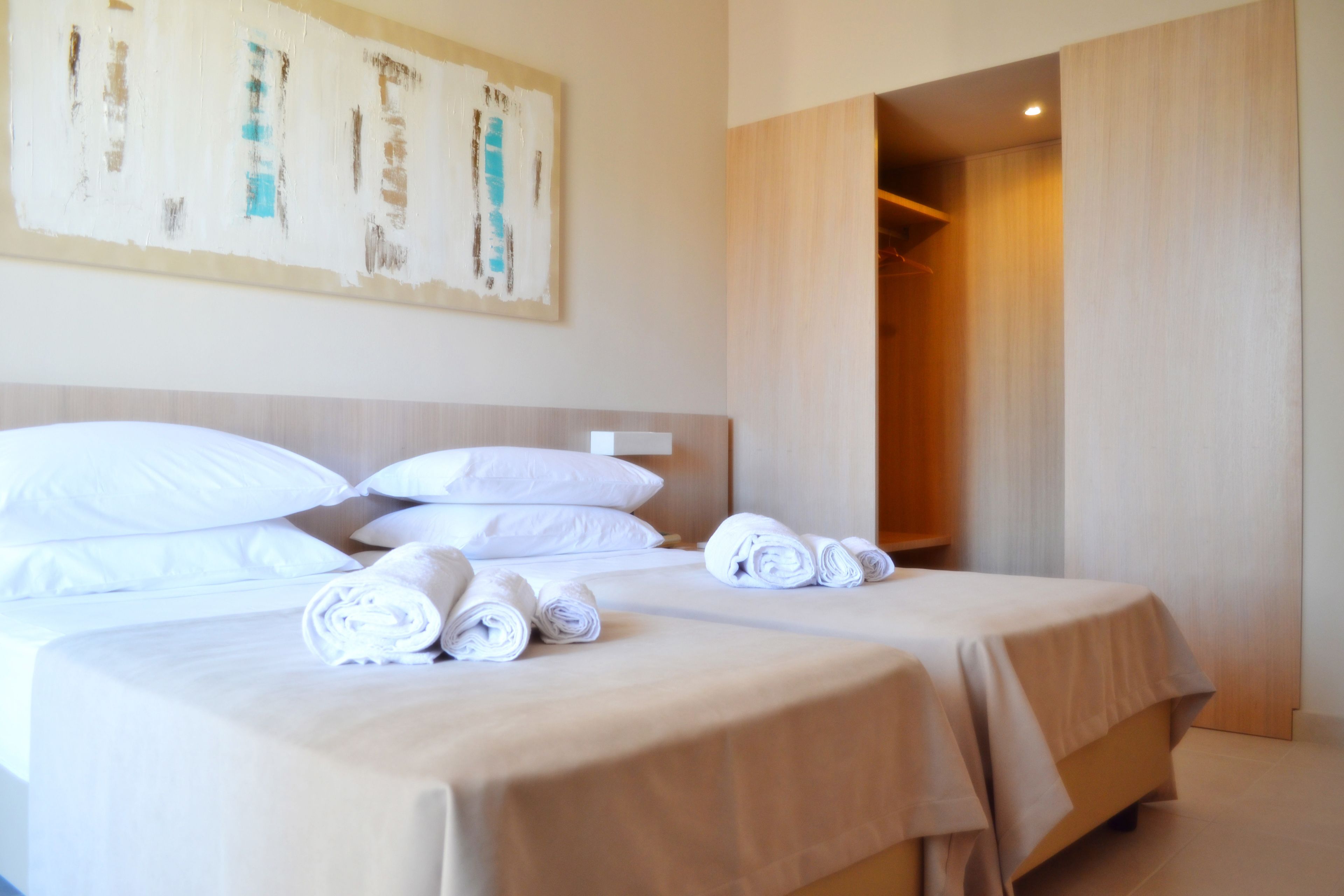 Een hotelkamer met twee bedden, elk voorzien van witte lakens en kussens, een schilderij aan de muur en een houten garderobe.