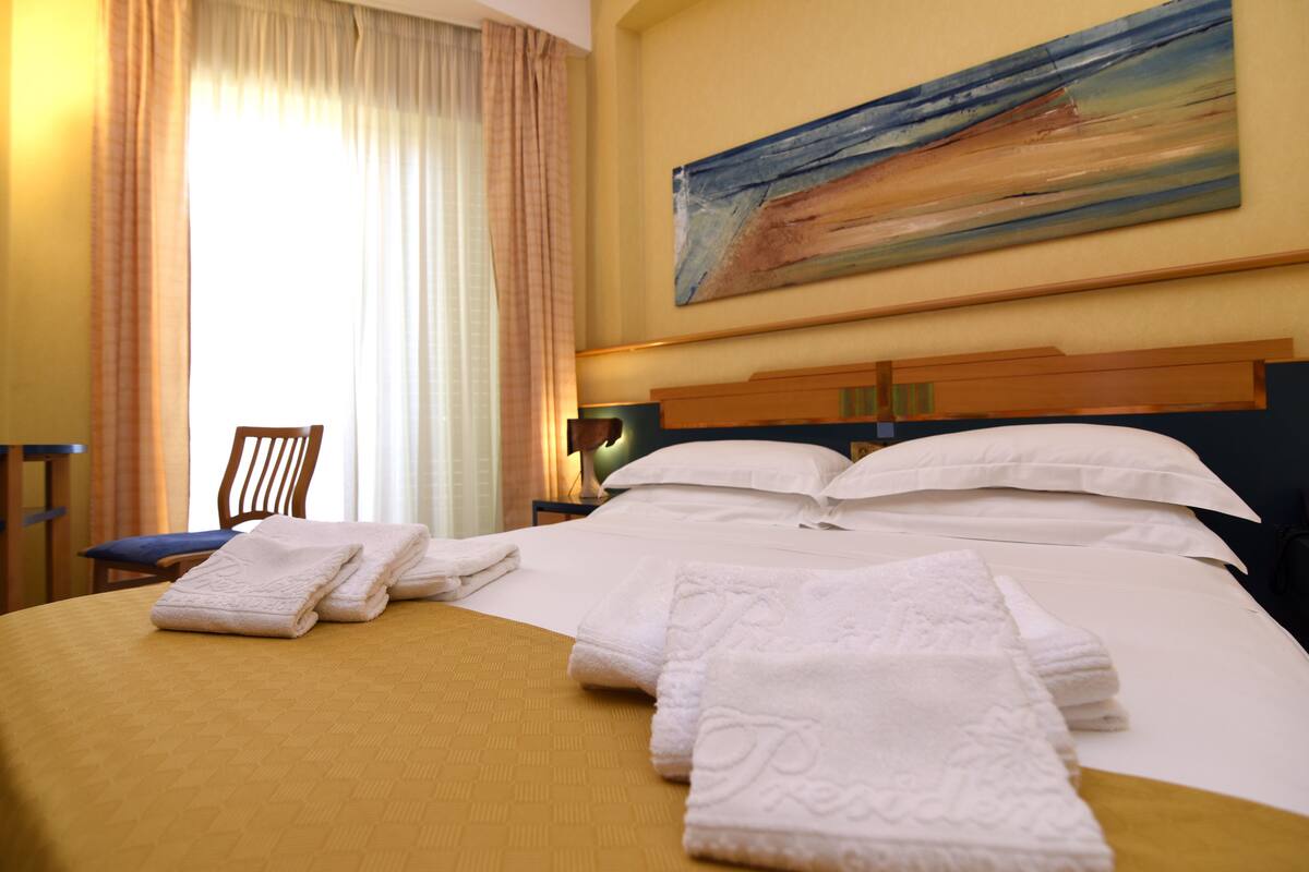 Ein Hotelzimmer mit einem großen Bett, ordentlich gefalteten weißen Handtüchern, einem Gemälde an der Wand und einem Sessel am Fenster.
