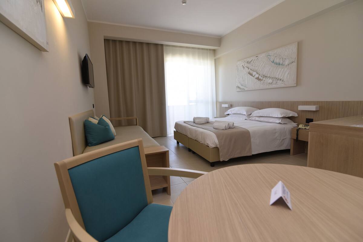 Ein Hotelzimmer mit Bett, Schreibtisch, Stuhl, Tisch, Fernseher und einem Fenster mit Vorhängen.