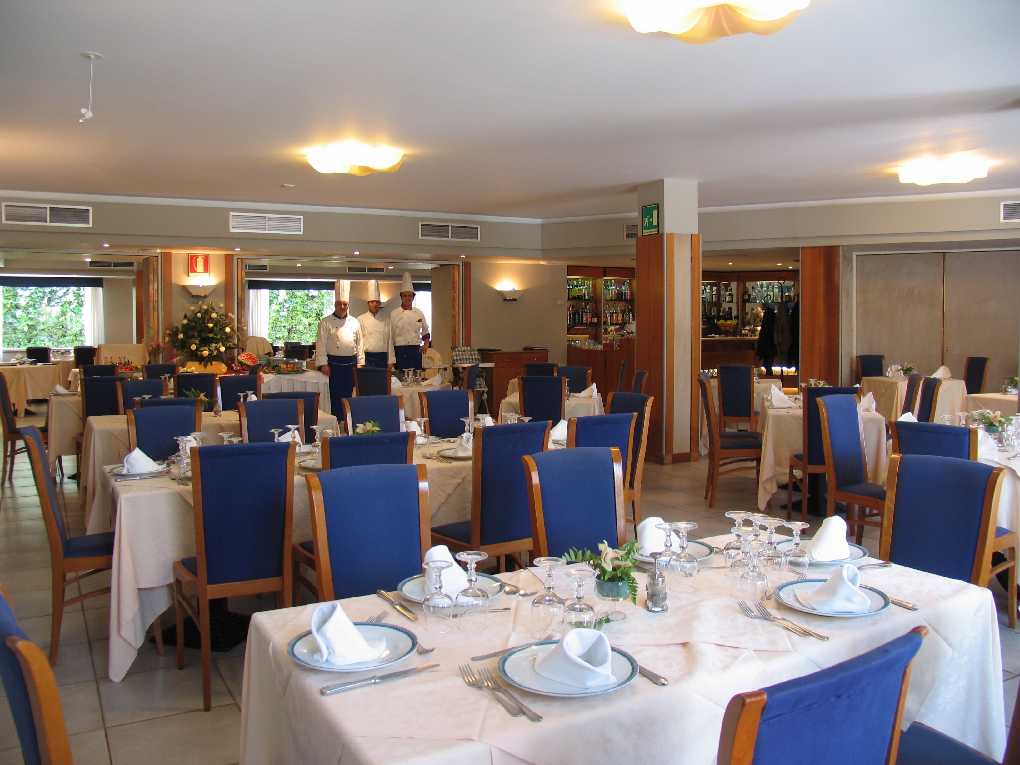 Restaurante com mesas preparadas para refeições, cadeiras azuis e chefs ao fundo.