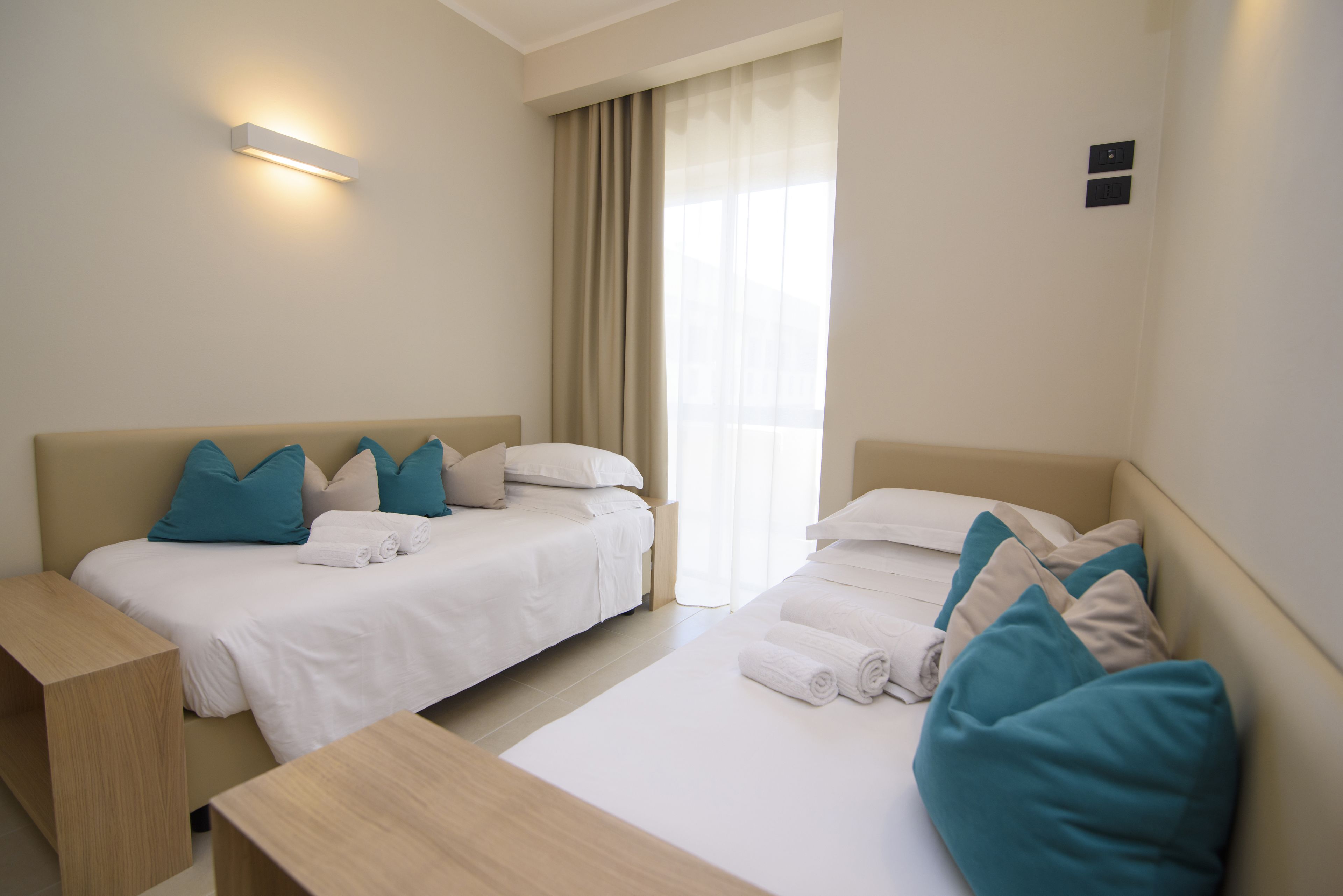 Habitación de hotel con dos camas, una mesita de noche de madera y un ventanal grande con cortinas.