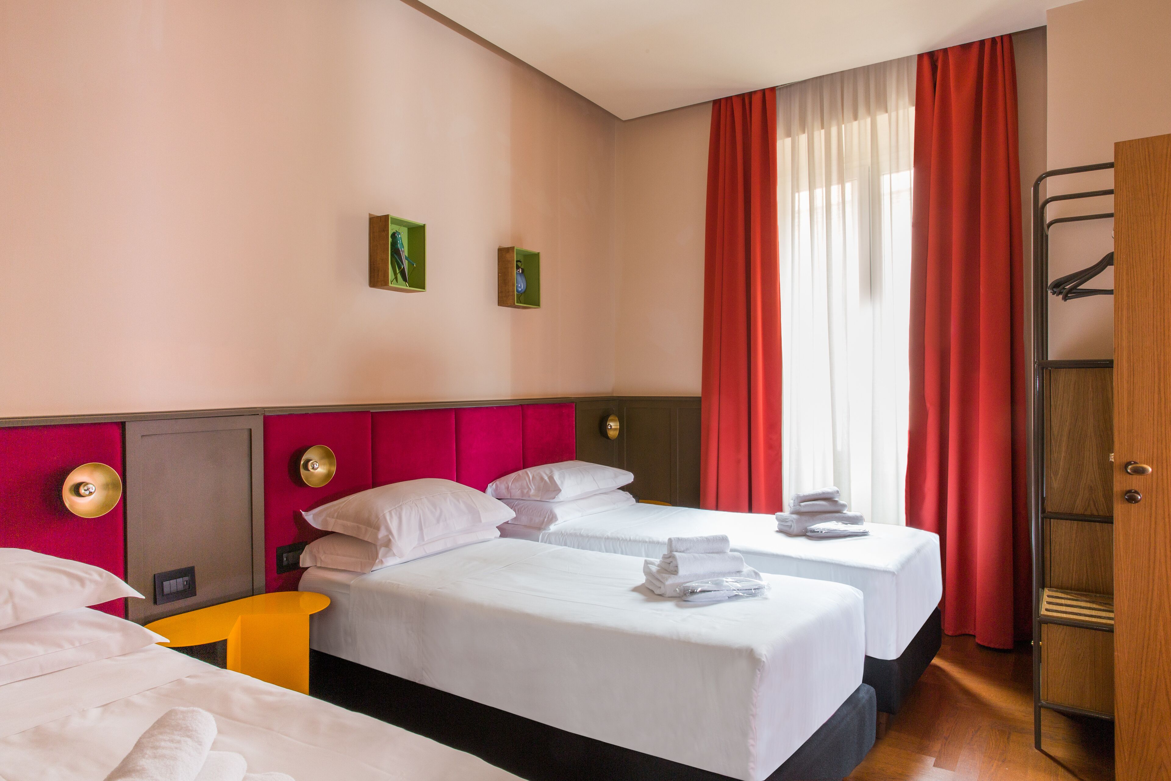 A hotel room with two beds, a wooden wardrobe, a small table, and a window with red curtains.