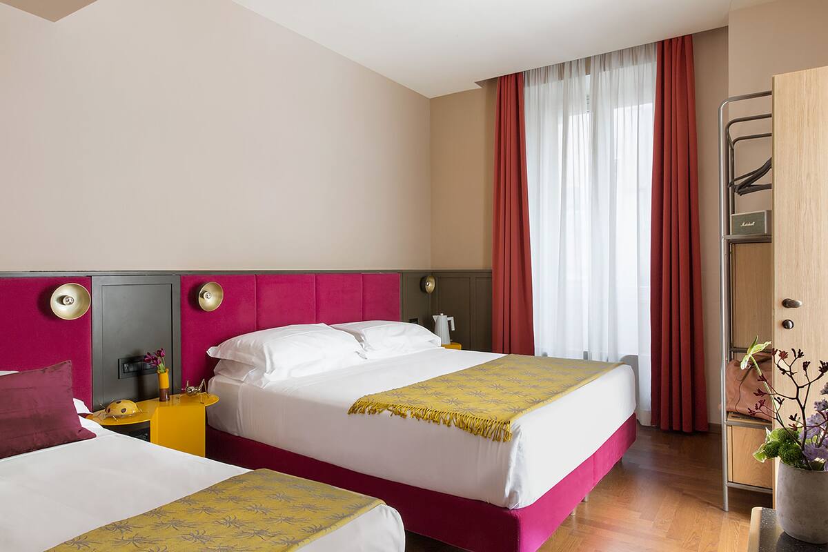 A hotel room with two beds, a wooden wardrobe, a nightstand with a lamp, and a window with curtains.