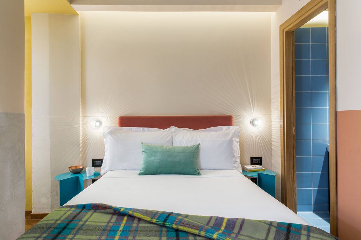 A neatly made bed with white linens, a green pillow, and a plaid blanket. There are two bedside tables with lamps, one on each side of the bed. The room has a wooden headboard and a blue tiled wall.