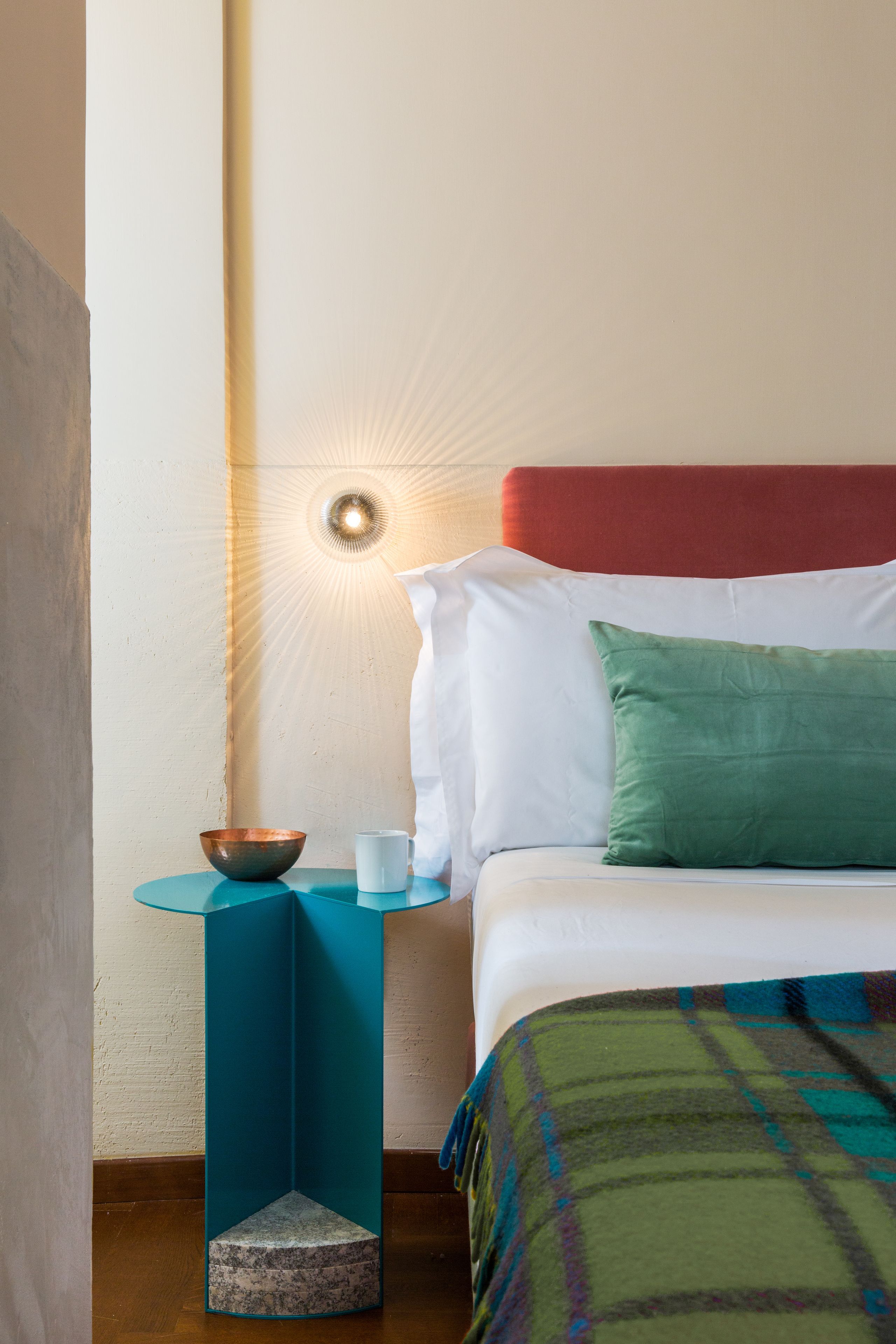 A neatly made bed with a red headboard, a green pillow, and a plaid blanket. A small table with a bowl and a white mug is beside the bed.