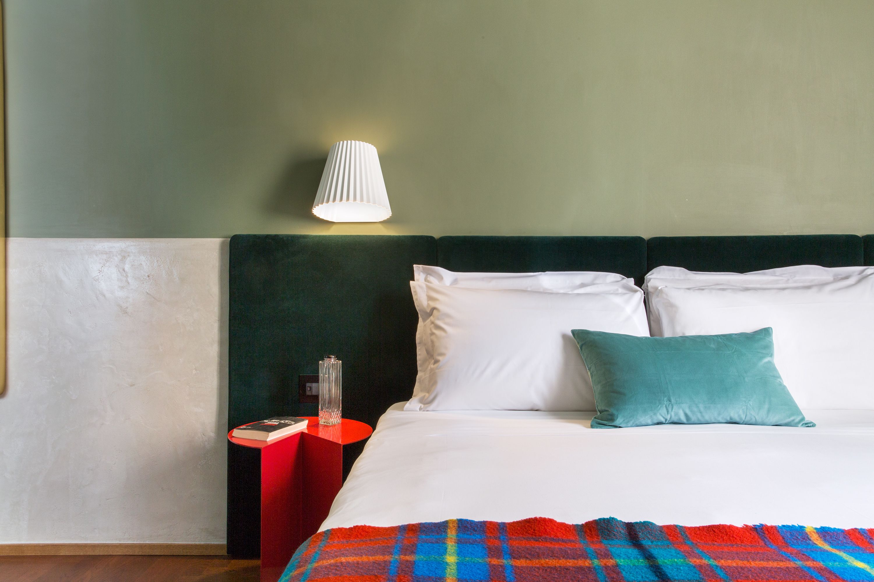 A hotel room with a bed, a nightstand, a lamp, and a red side table.