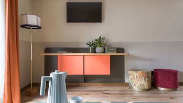 Een moderne woonkamer met een flatscreen televisie aan de muur, een houten vloer, een blauwe yogamat, een wit boek, een hoge vloerlamp, een klein tafeltje met een bloevas, en twee decoratieve krukken.