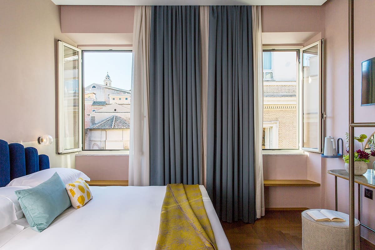 A hotel room with a bed, a window with curtains, a small table with a vase, and a view of buildings outside.