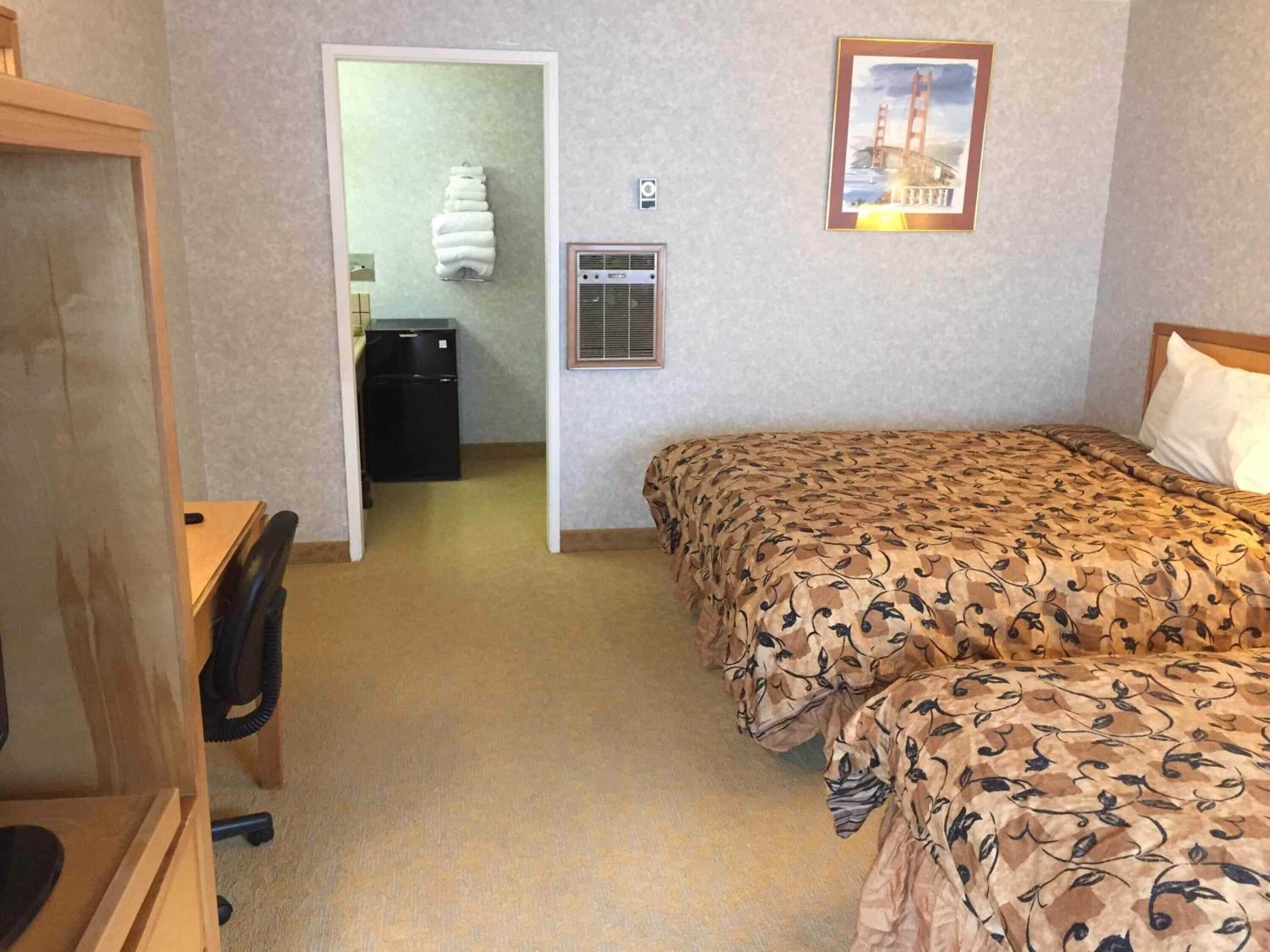 A hotel room with two beds, a desk, a chair, a small refrigerator, a wall-mounted heater, and a framed picture of a bridge.