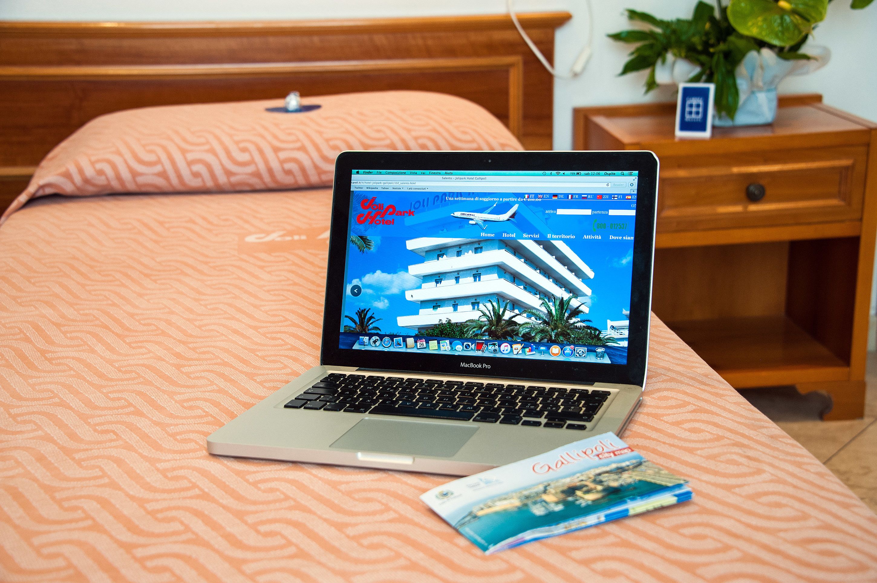 A MacBook Pro on a bed with a patterned bedspread, a magazine, and a wooden nightstand with a plant.