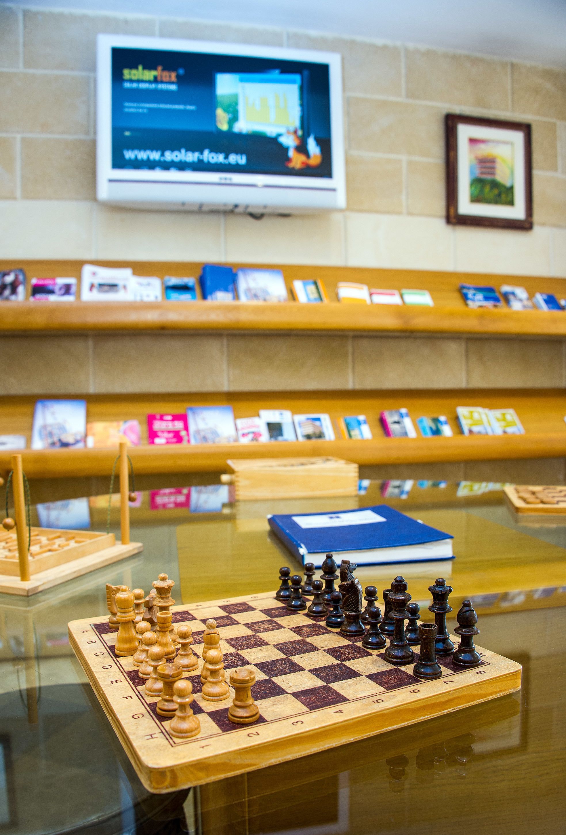 A chessboard with pieces on a glass table, surrounded by bookshelves with books and a framed picture on the wall.