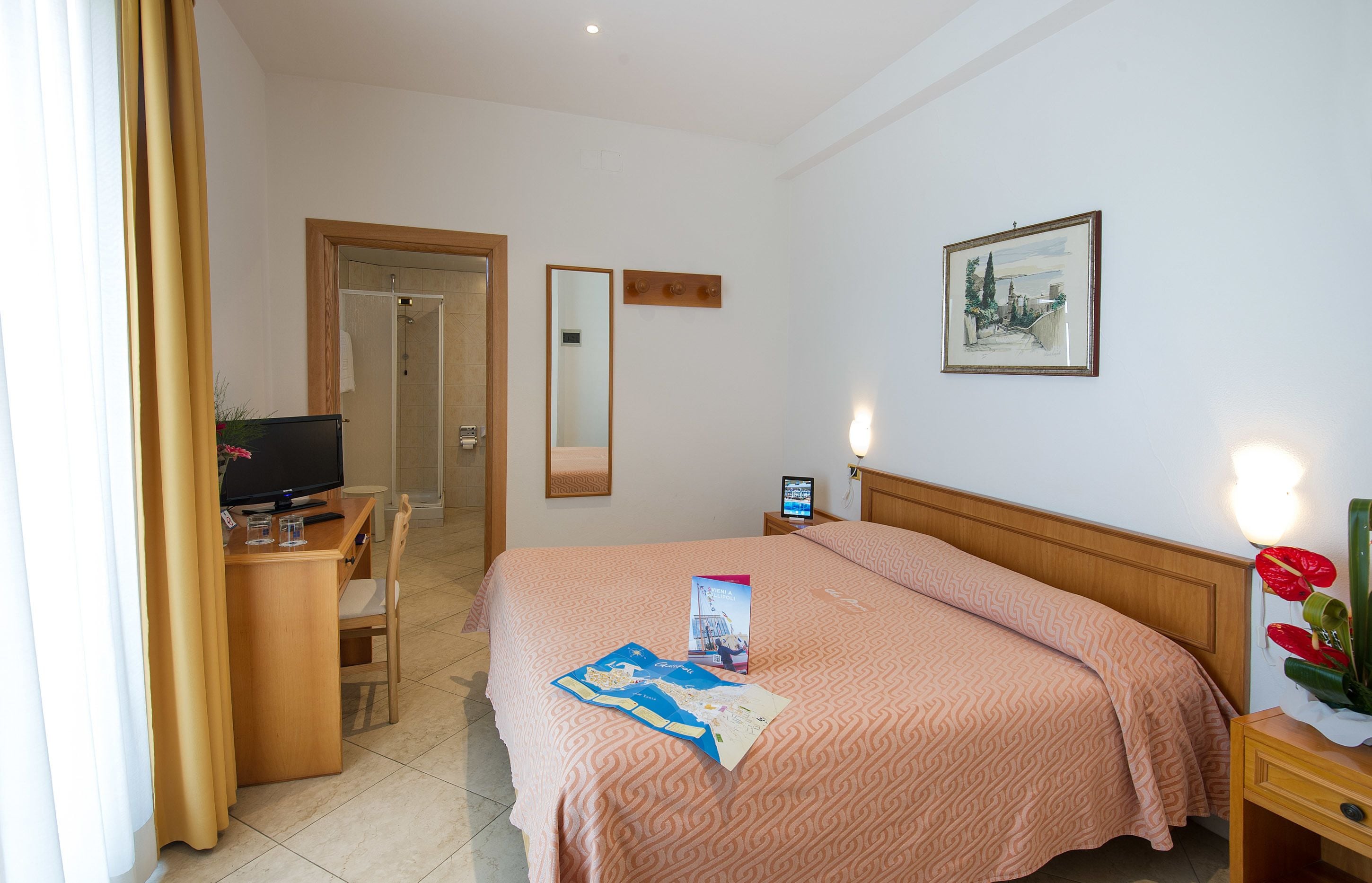 Habitación de hotel con cama, escritorio con computadora, silla, televisor, espejo y un cuadro en la pared.