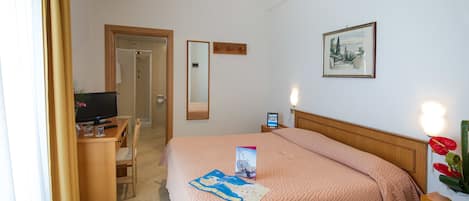 Ett hotellrum med en säng, ett skrivbord med en dator, en stol, en tv, en spegel och en inramad tavla på väggen.