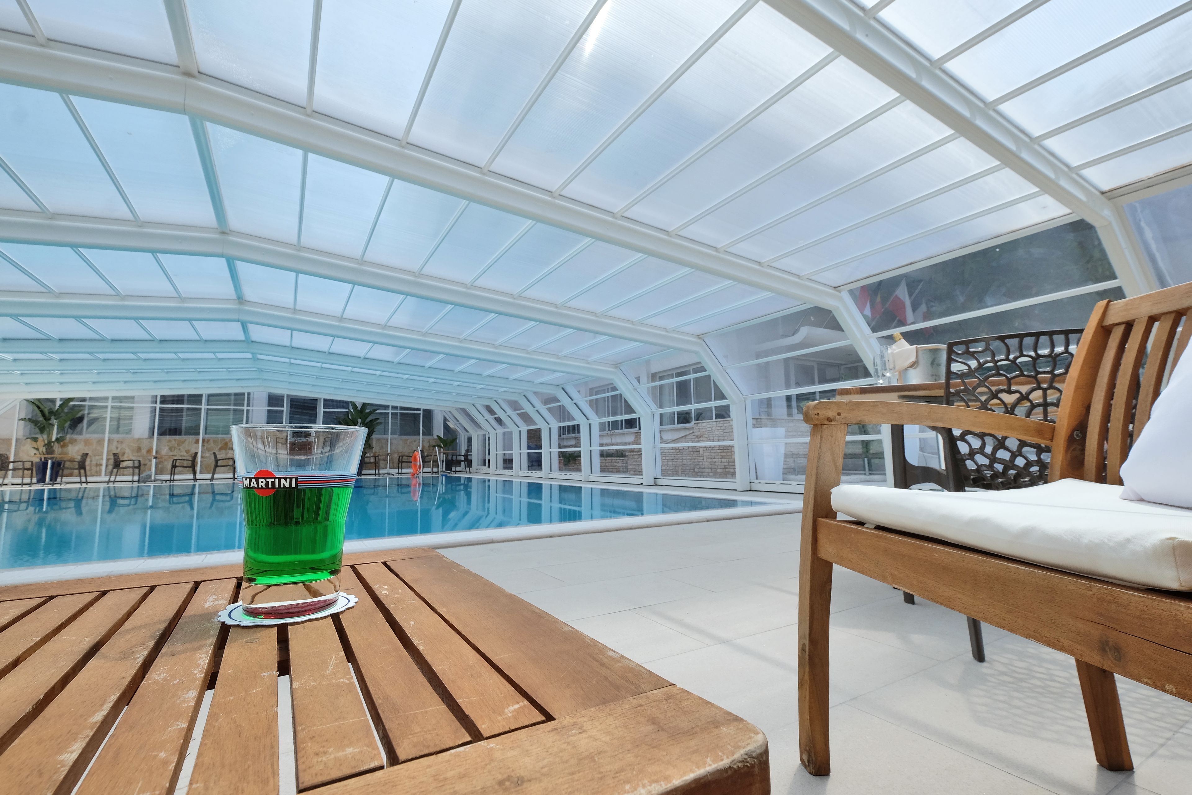 A glass of Martini on a wooden table by a pool in a glasshouse structure.