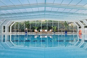 An indoor swimming pool with clear glass walls, wooden lounge chairs, and potted plants.