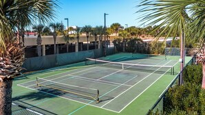 Una cancha de tenis con red, rodeada de palmeras y un edificio al fondo.