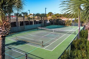Un court de tennis avec un filet, entouré de palmiers et avec un bâtiment en arrière-plan.
