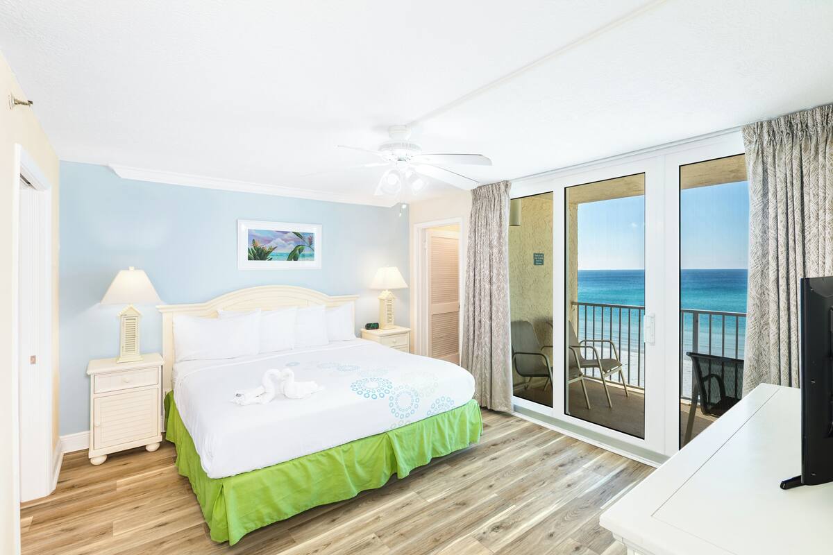 A hotel room with a bed, two bedside tables, a ceiling fan, a view of the ocean through a sliding door, and a TV on a stand.