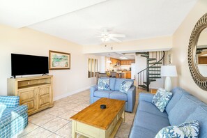 Un salon avec un téléviseur, un canapé et une table basse. Un escalier mène à la cuisine.
