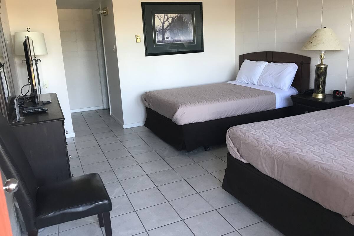 A hotel room with two beds, a desk with a lamp, a chair, and a framed picture on the wall.