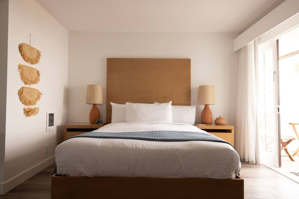 Ein modernes Schlafzimmer mit einem großen Bett, zwei Nachttischen mit Lampen, einem hölzernen Kopfteil und Blick ins Freie durch eine Schiebetür.