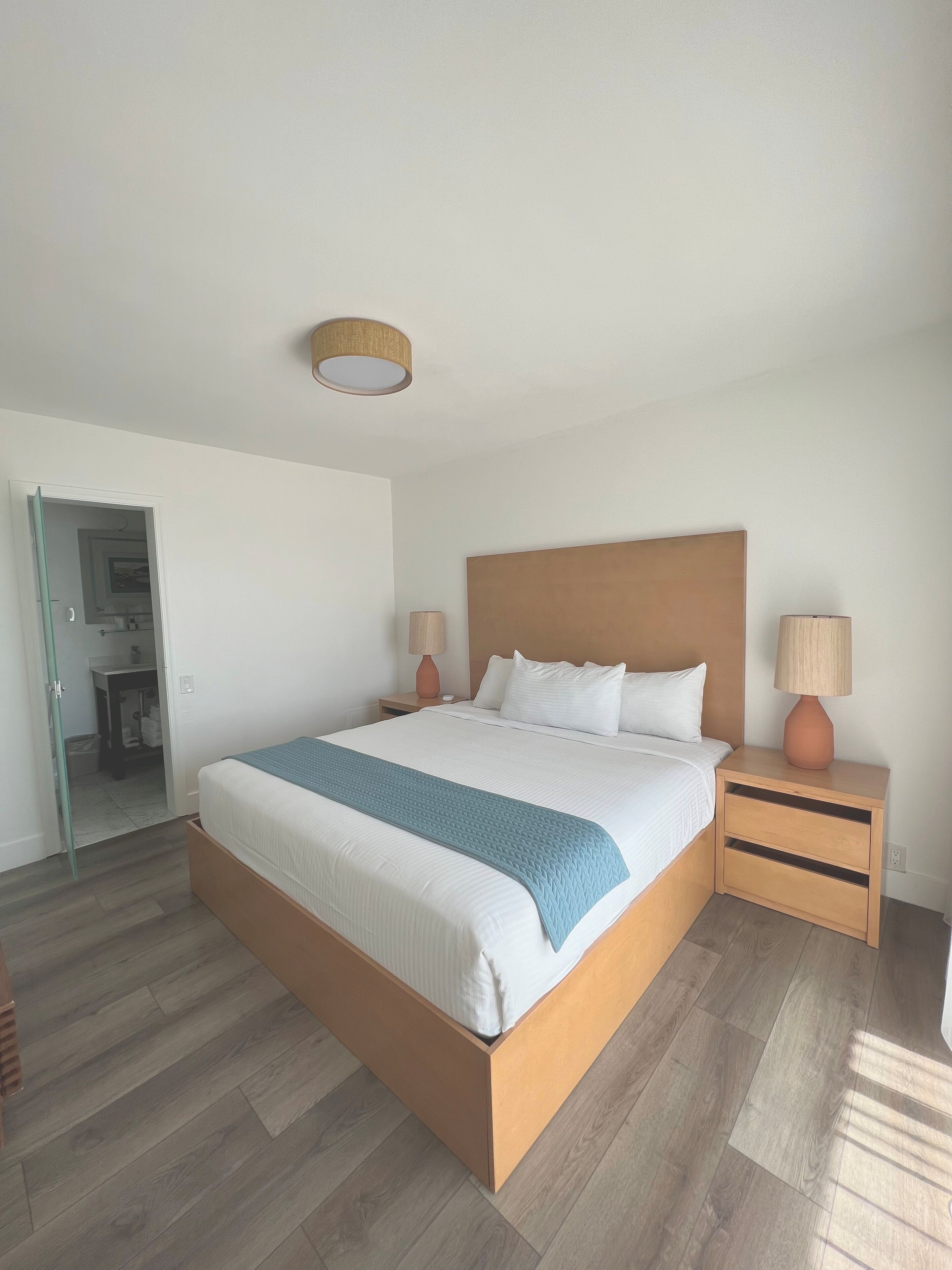 Habitación de hotel con una cama grande, cabecero de madera, dos mesitas de noche con lámparas y vistas a un baño a través de una puerta abierta.