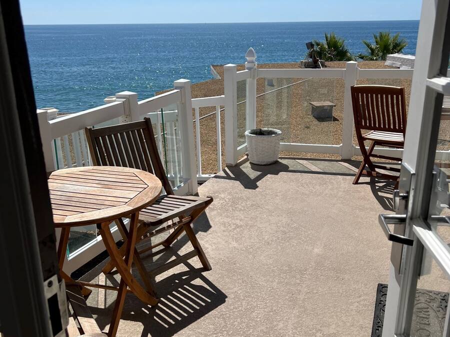 Ein Balkon mit einem runden Holztisch und zwei Holzstühlen, mit Blick auf einen Strand und das Meer im Hintergrund.