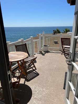 Un balcón con una mesa redonda de madera y dos sillas de madera, con vista a una playa y el océano al fondo.