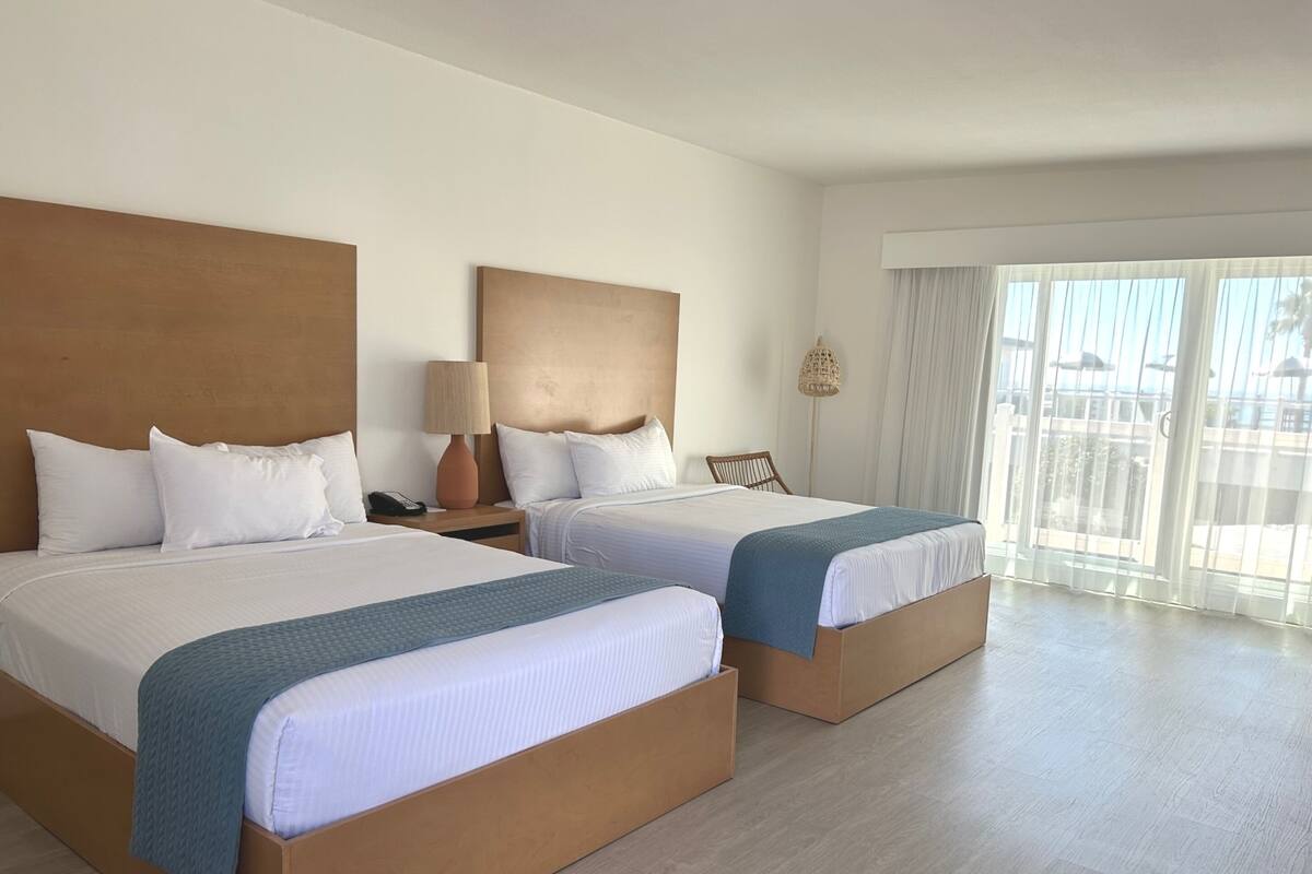 Ein Hotelzimmer mit zwei Betten, einem Nachttisch mit Lampe, einem Sessel und einem großen Fenster mit Vorhängen.