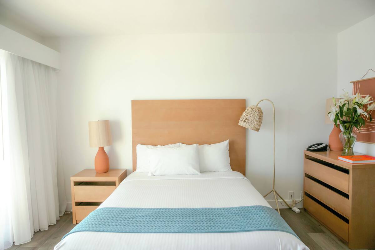 Ein ordentlich eingerichtetes Hotelzimmer mit Bett, Nachttisch, Lampe, Kommode und einer Vase mit Blumen.