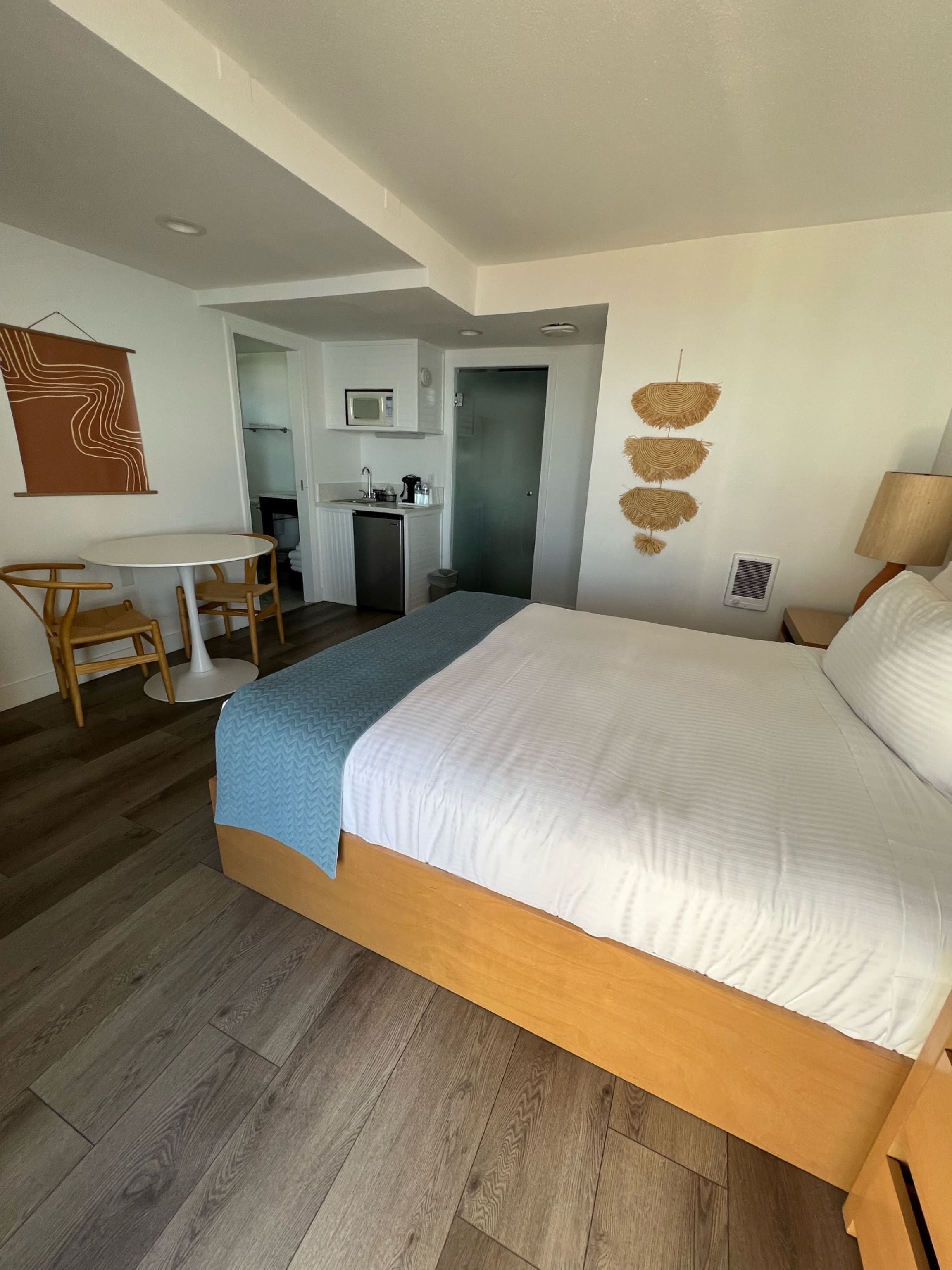 A hotel room with a large bed, a desk with chairs, a kitchenette, and a bathroom visible through an open door.