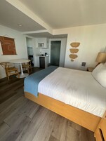 A hotel room with a large bed, a desk with chairs, a kitchenette, and a bathroom visible through an open door.