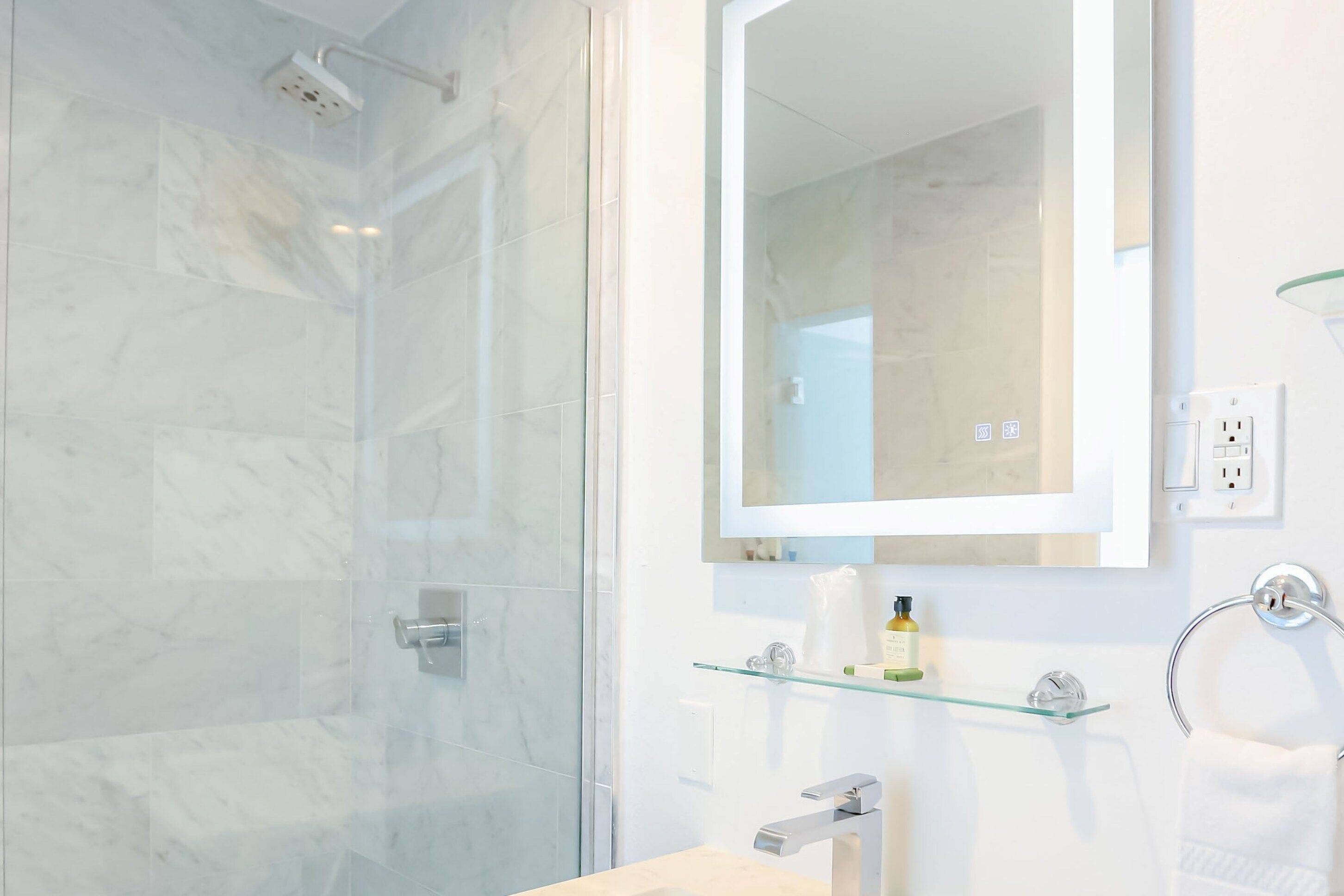 Un bagno moderno con cabina doccia in vetro, un lavandino bianco, una mensola in vetro con articoli da toilette e uno specchio grande.