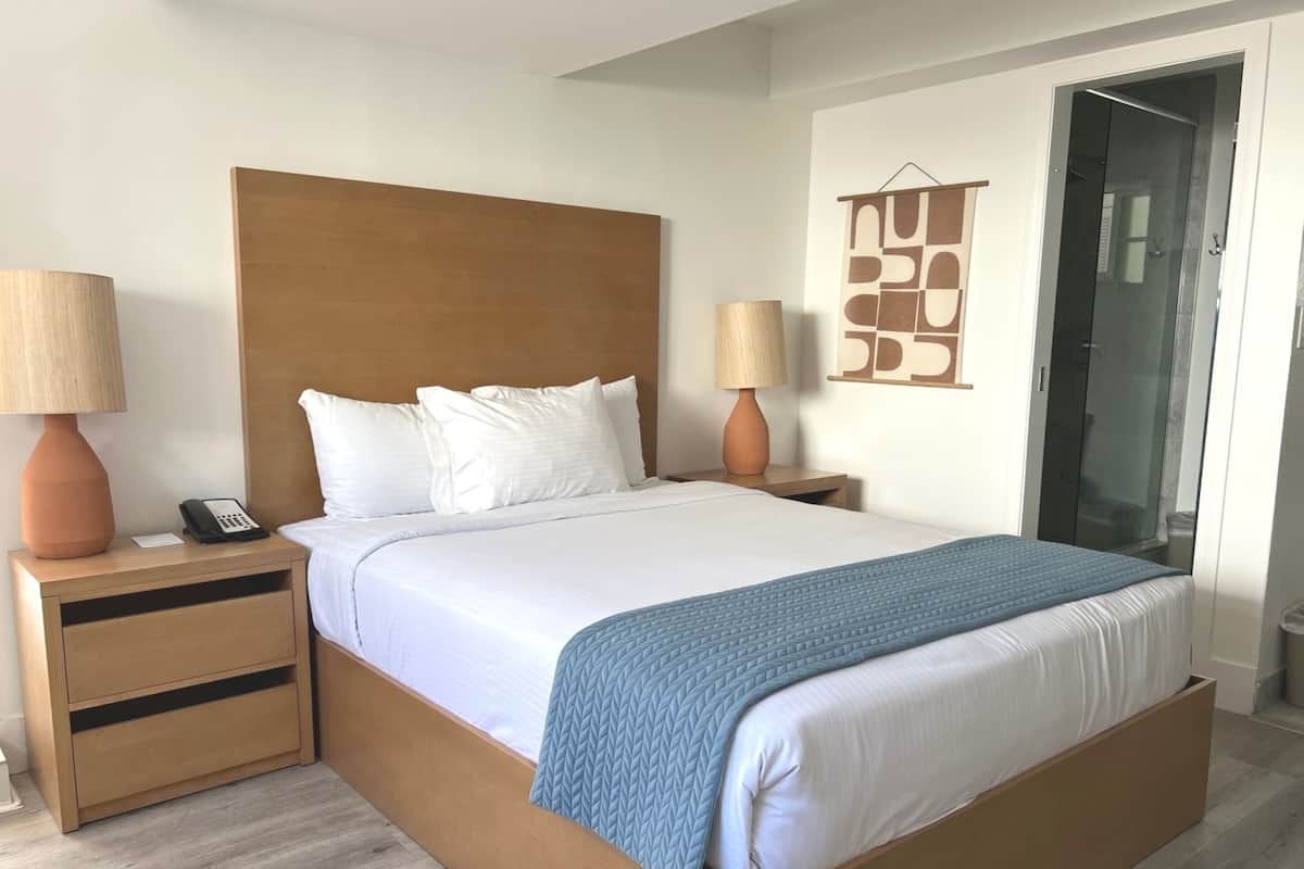 Ein modernes Hotelzimmer mit einem hölzernen Bettgestell, weißer Bettwäsche, zwei Nachttischen mit Lampen, einem Telefon und einem an der Wand hängenden Kunstwerk.