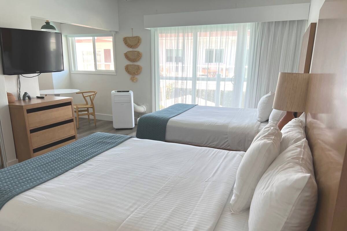 Ein Hotelzimmer mit zwei Betten, einem Schreibtisch, einem Stuhl, einem Fernseher und einem Fenster mit Vorhängen.