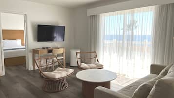 A hotel room with a bed, a TV, a round table, two wicker chairs, a sofa, and a large window with a view of the sea.