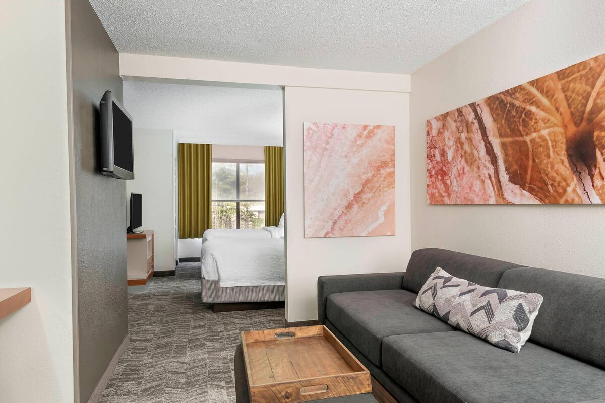 Habitación de hotel con sofá gris, una mesita de centro de madera, cama con edredón blanco y televisor montado en la pared.