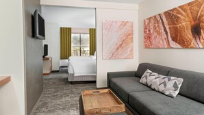 Habitación de hotel con sofá gris, una mesita de centro de madera, cama con edredón blanco y televisor montado en la pared.
